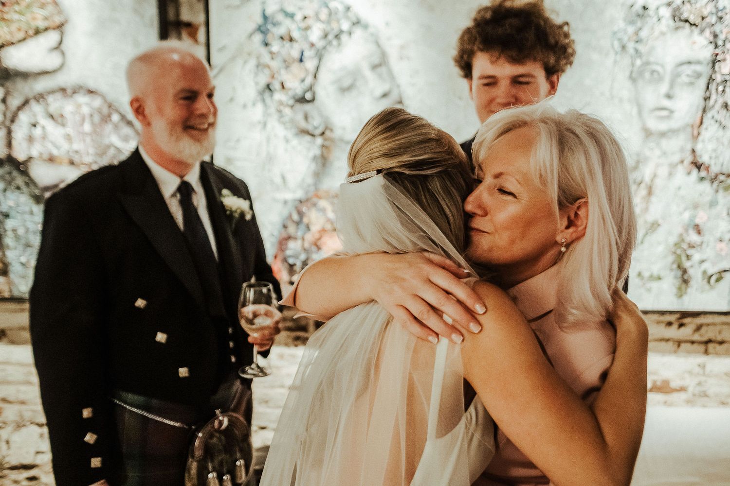 Warm wedding reception moment with guests embracing bride in venue with artistic wall decor.