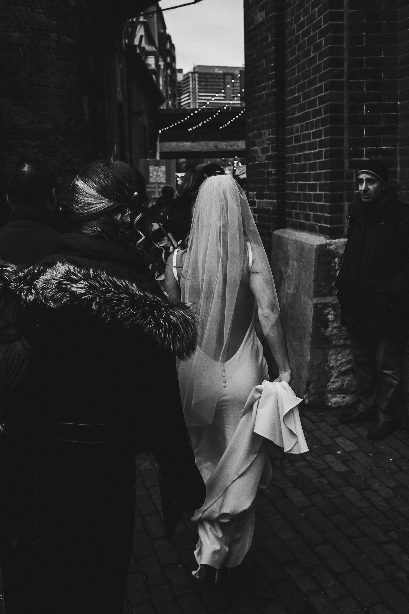 Bride in form-fitting dress and veil walking through urban alley at night.