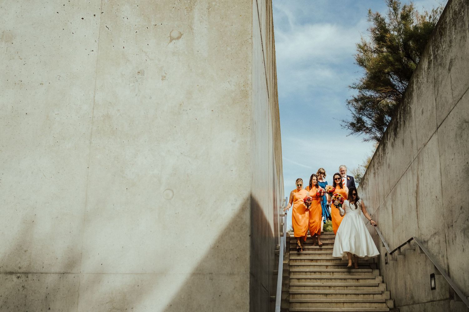 Wedding party in vibrant orange and white attire stands on concrete steps against a bright sky.