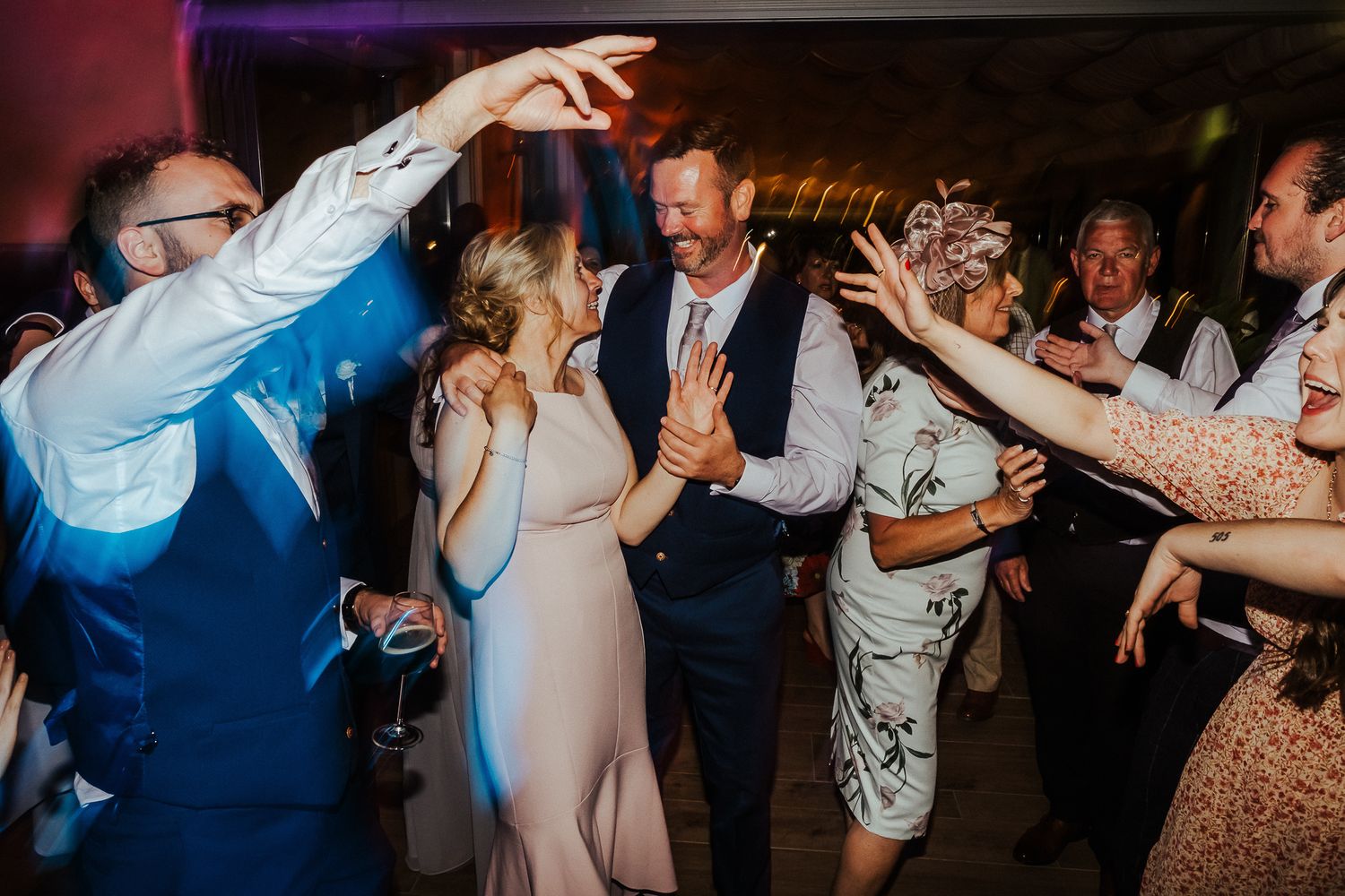 Wedding guests dancing enthusiastically on a crowded dance floor with blue and red lighting.