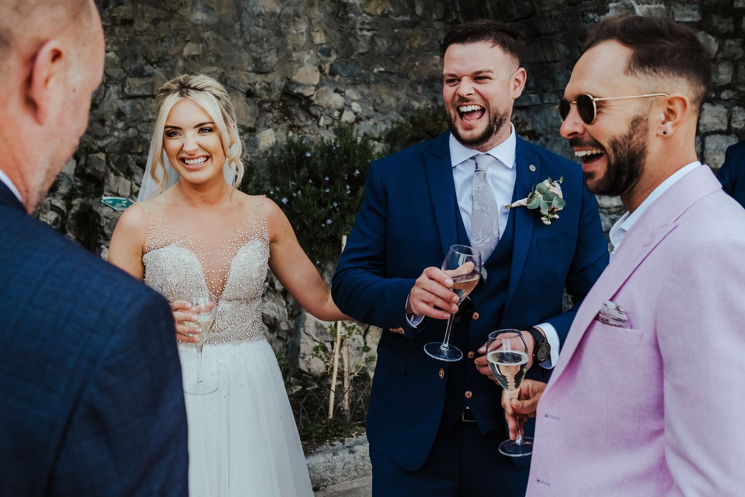 Wedding party enjoying celebratory drinks against a stone wall backdrop.