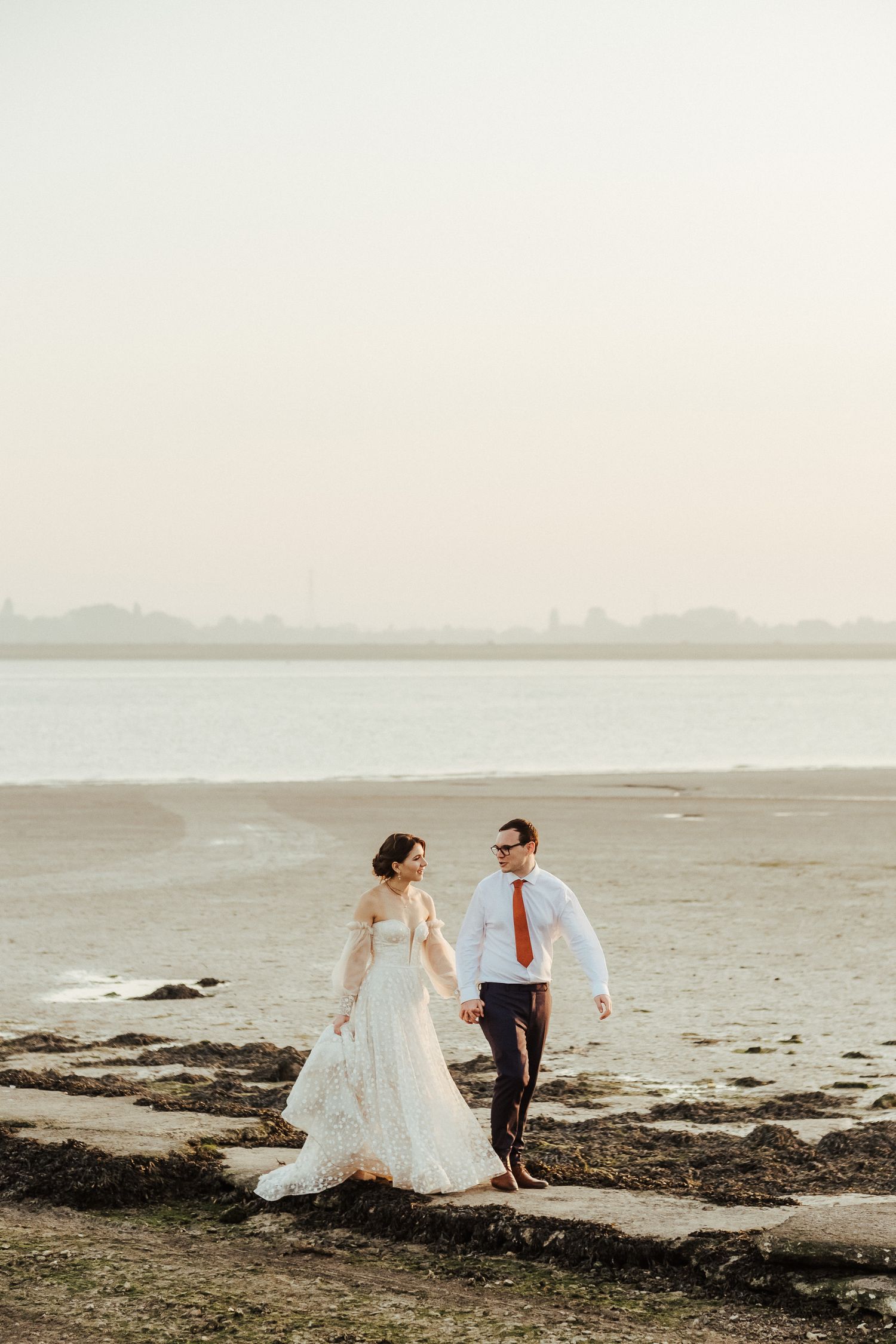 A couple walks hand in hand along a deserted beach at sunset in wedding attire.