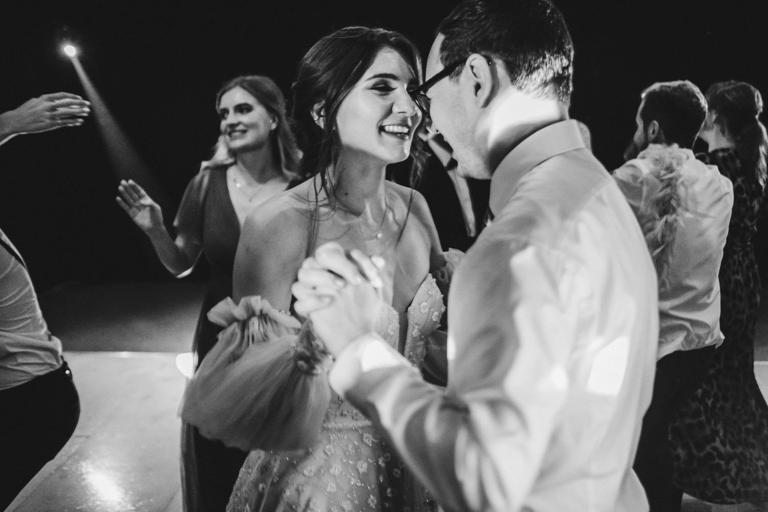 Wedding couple dances together while guests celebrate on dimly lit dance floor.