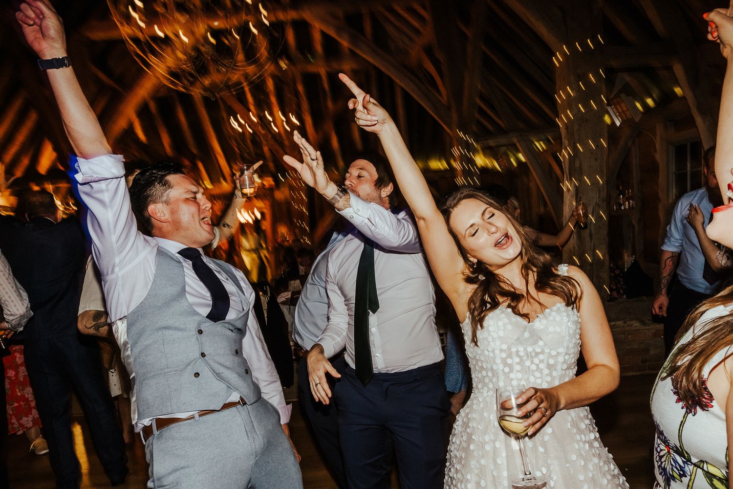 Wedding guests dancing energetically at a reception with wooden beams overhead.