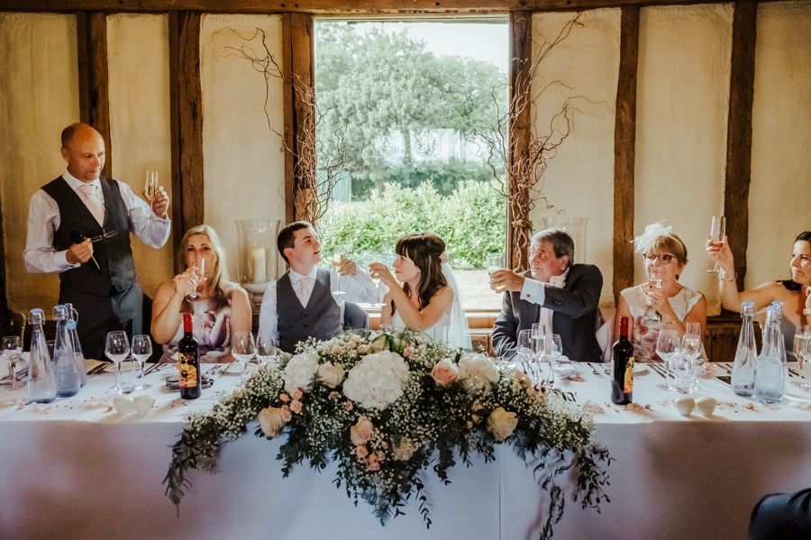 Long banquet table setup in rustic barn venue with floral decorations and window light.