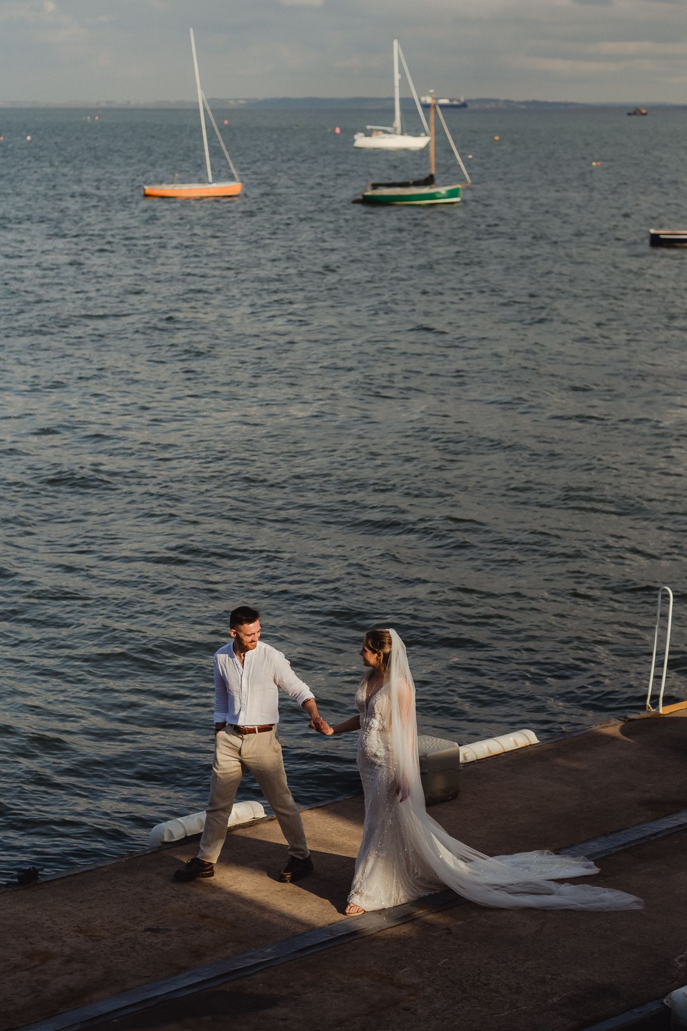 Two people in wedding attire hold hands while walking along a seaside dock with sailboats in the background.
