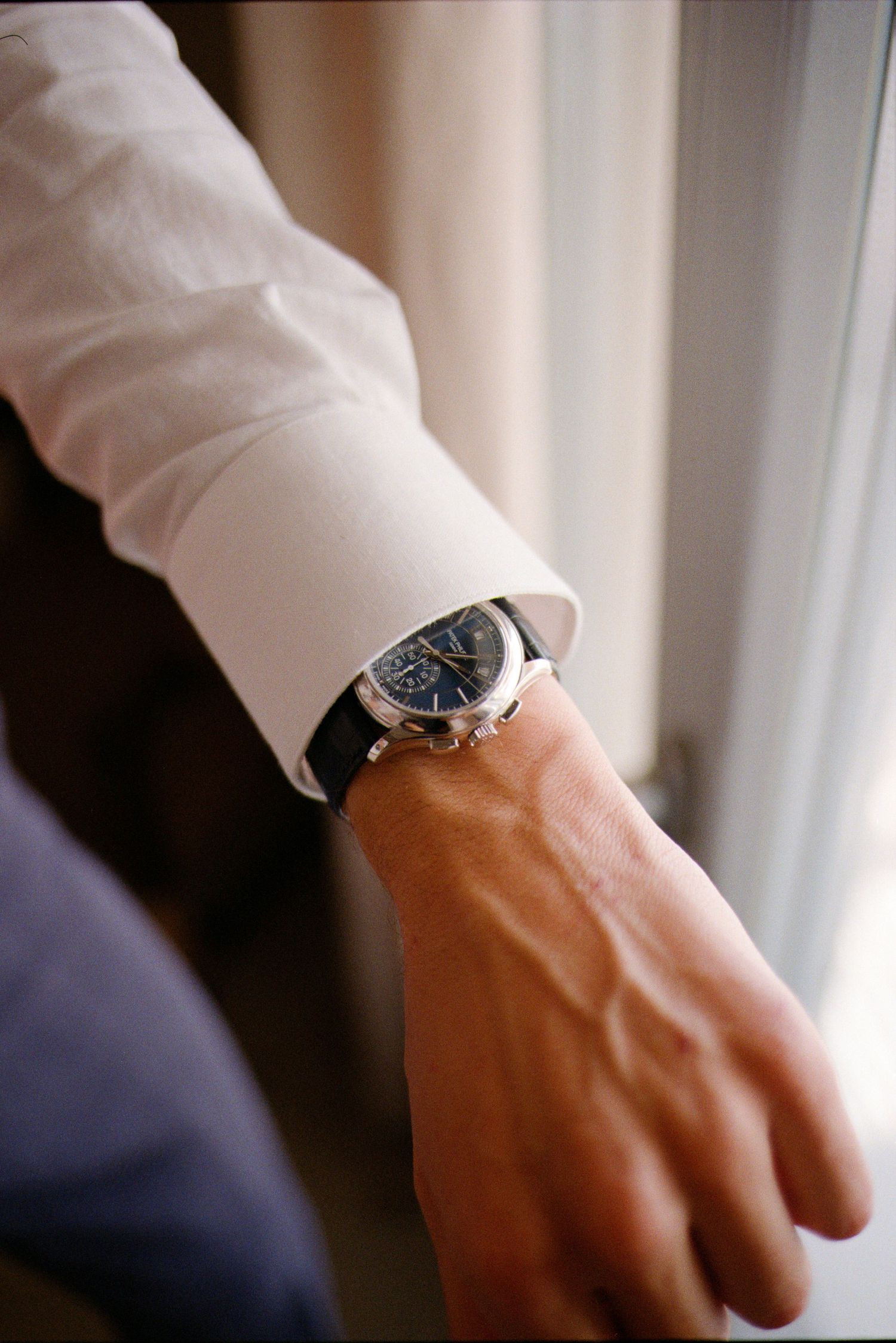 A watch-wearing hand adjusts a white dress shirt sleeve against a soft blurred background.