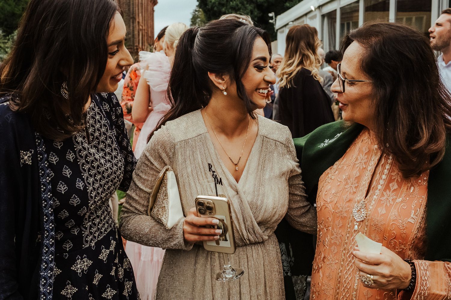 Three friends sharing laughs and drinks at an outdoor social gathering while wearing casual evening attire.