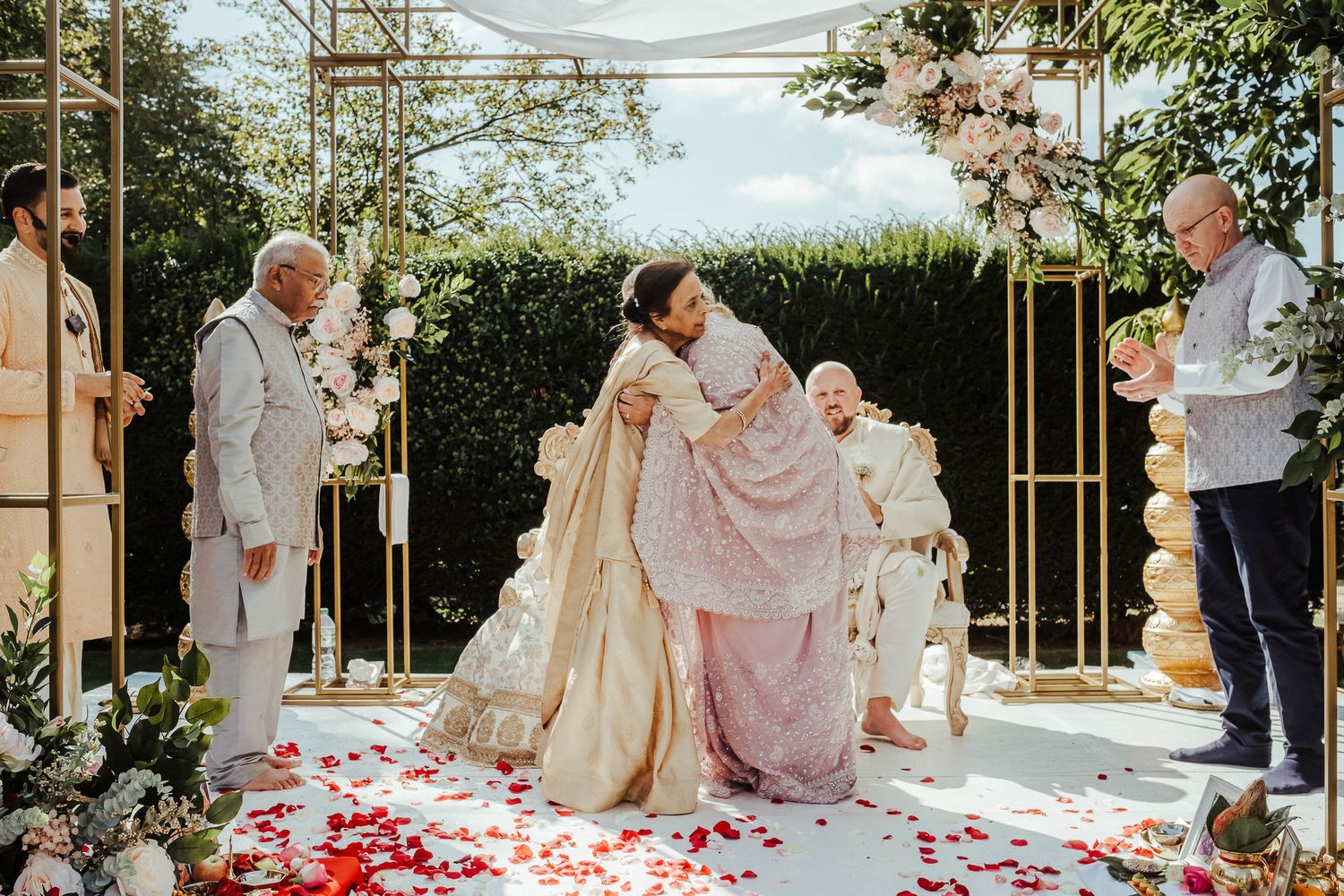 An outdoor wedding ceremony with rose petals scattered on white carpet under a floral arch with gold accents.
