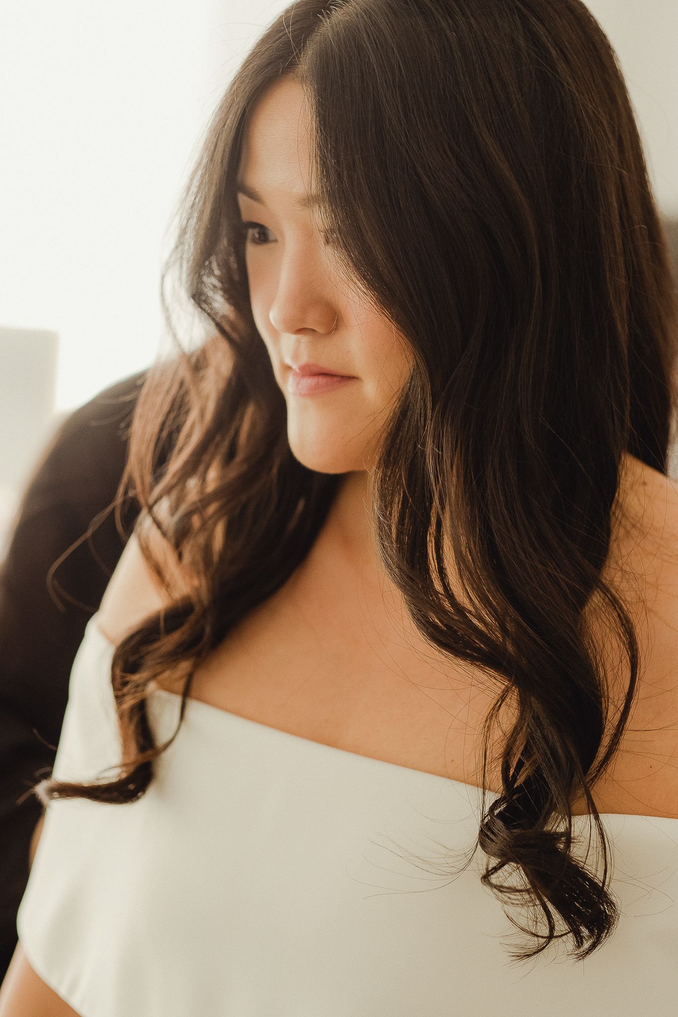 Person with long dark wavy hair wearing white off-shoulder top poses near window in soft natural lighting.