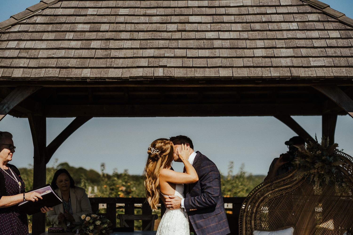 A romantic wedding ceremony takes place under a wooden gazebo on a bright summer day.
