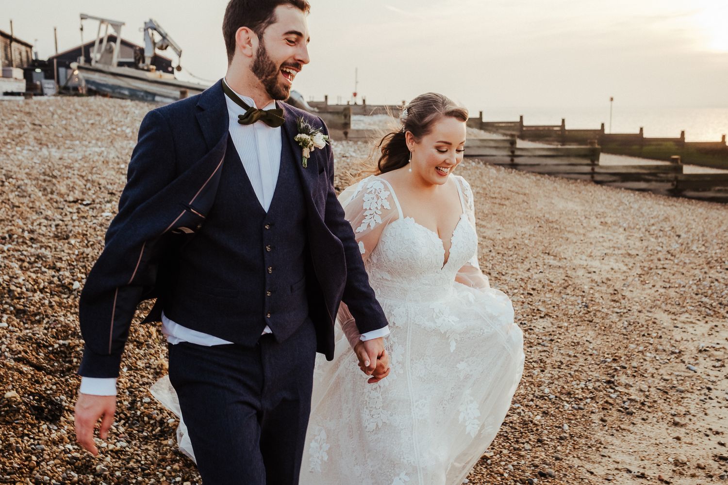 Couple walks joyfully along a beach boardwalk in full wedding attire.