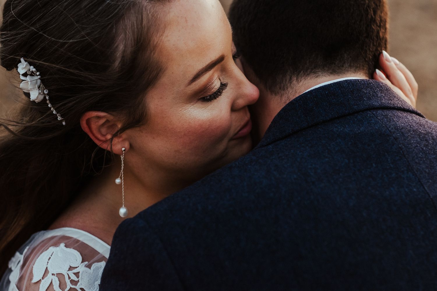 Close-up of a bride with elegant makeup and jewelry embracing her groom from behind.