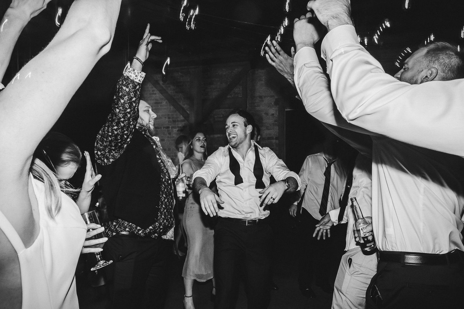 Black and white photo of wedding guests dancing with arms raised during an enthusiastic celebration.