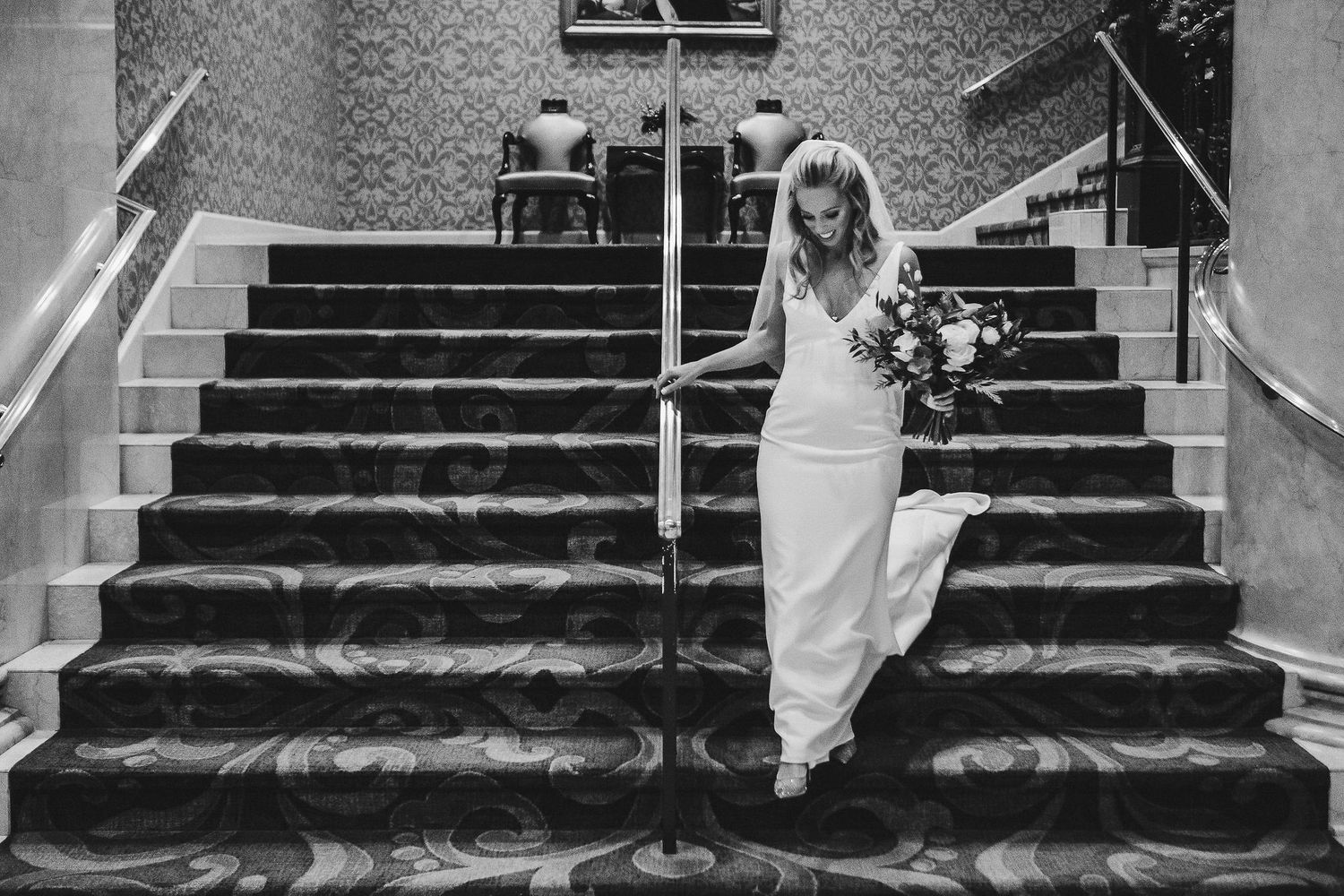 Bride in fitted white dress descending ornate staircase while holding bouquet.