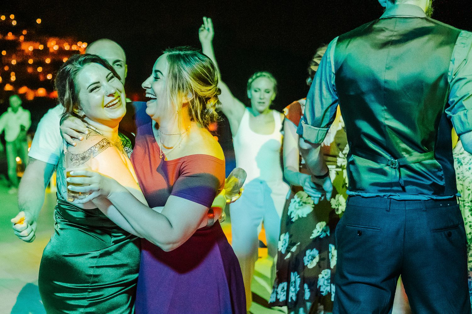 Wedding guests dancing and embracing under colorful green lighting at an evening reception.