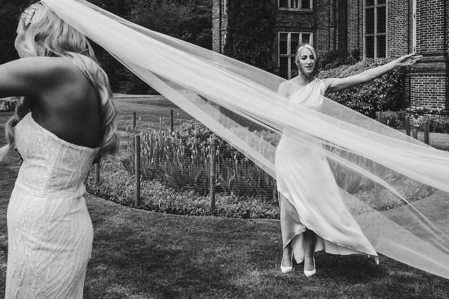 Black and white photo of playful moment with flowing wedding veil in garden setting.