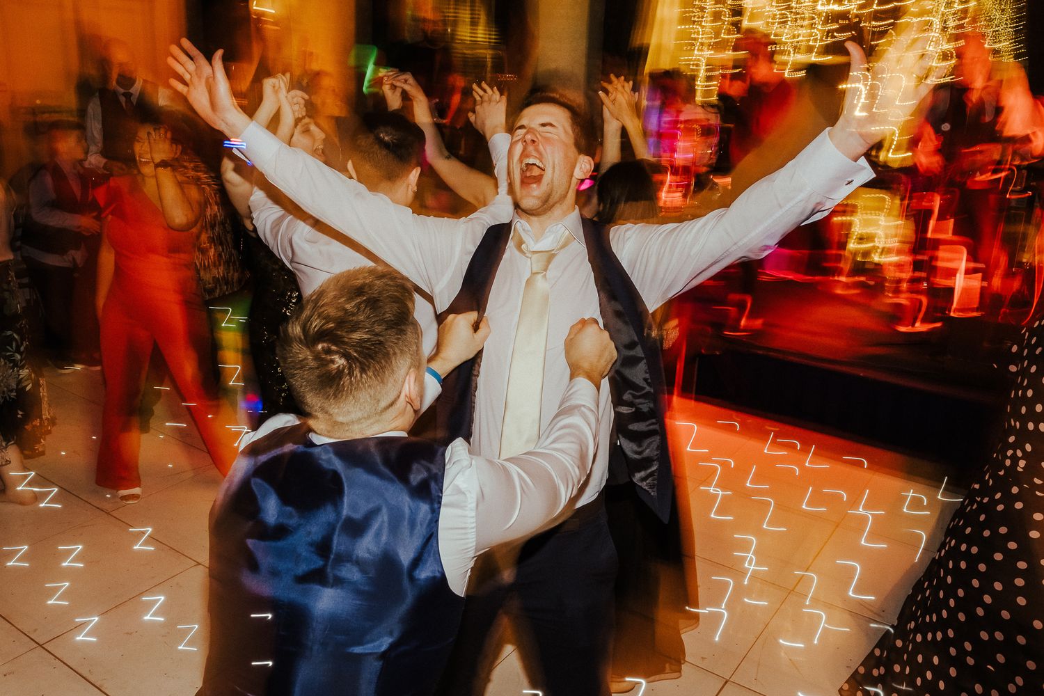 Person celebrating enthusiastically on dance floor with arms raised in colorful lighting.