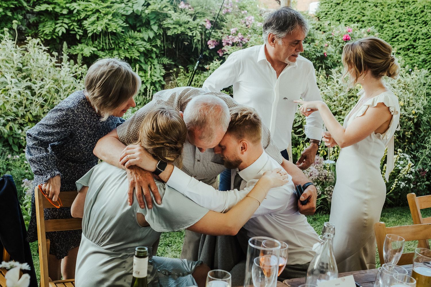 Wedding guests share an intimate emotional group hug at an outdoor garden reception.