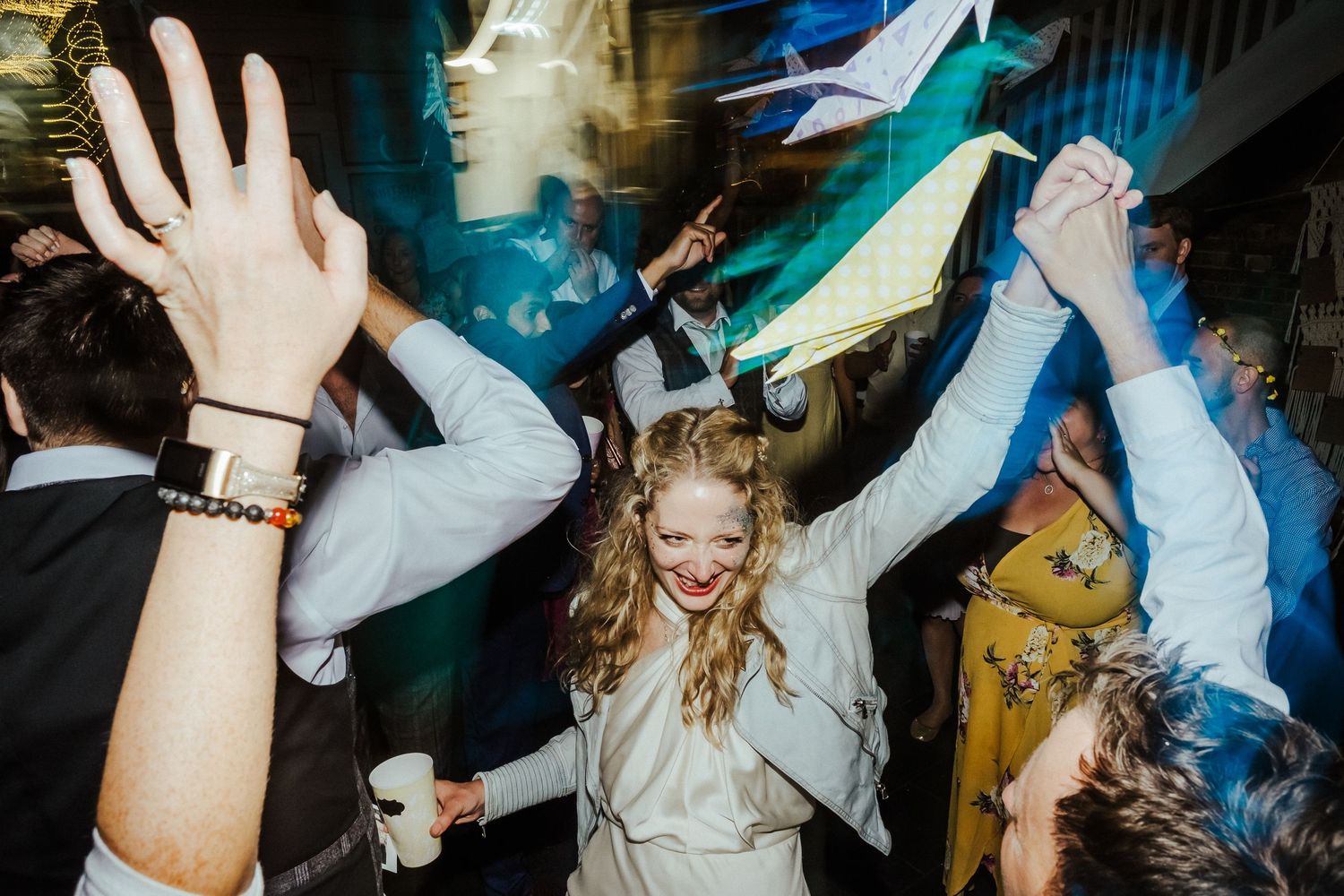 Wedding party dancing and celebrating with raised hands under decorative lighting.