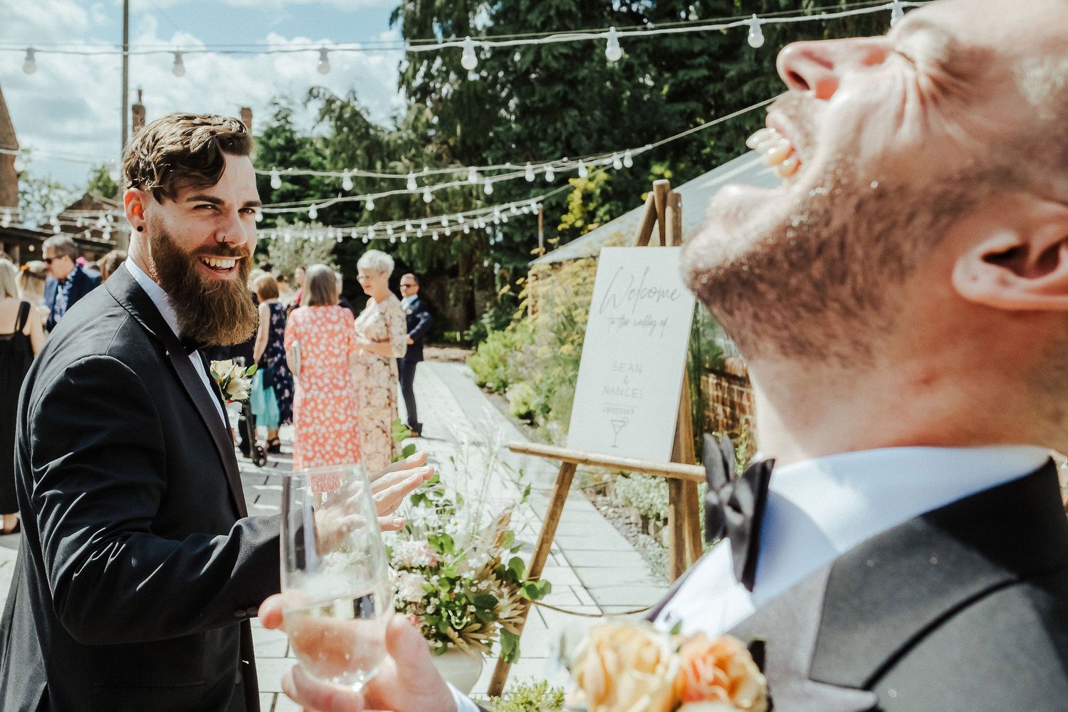 Party guests enjoying drinks and laughter at an outdoor wedding reception with string lights.