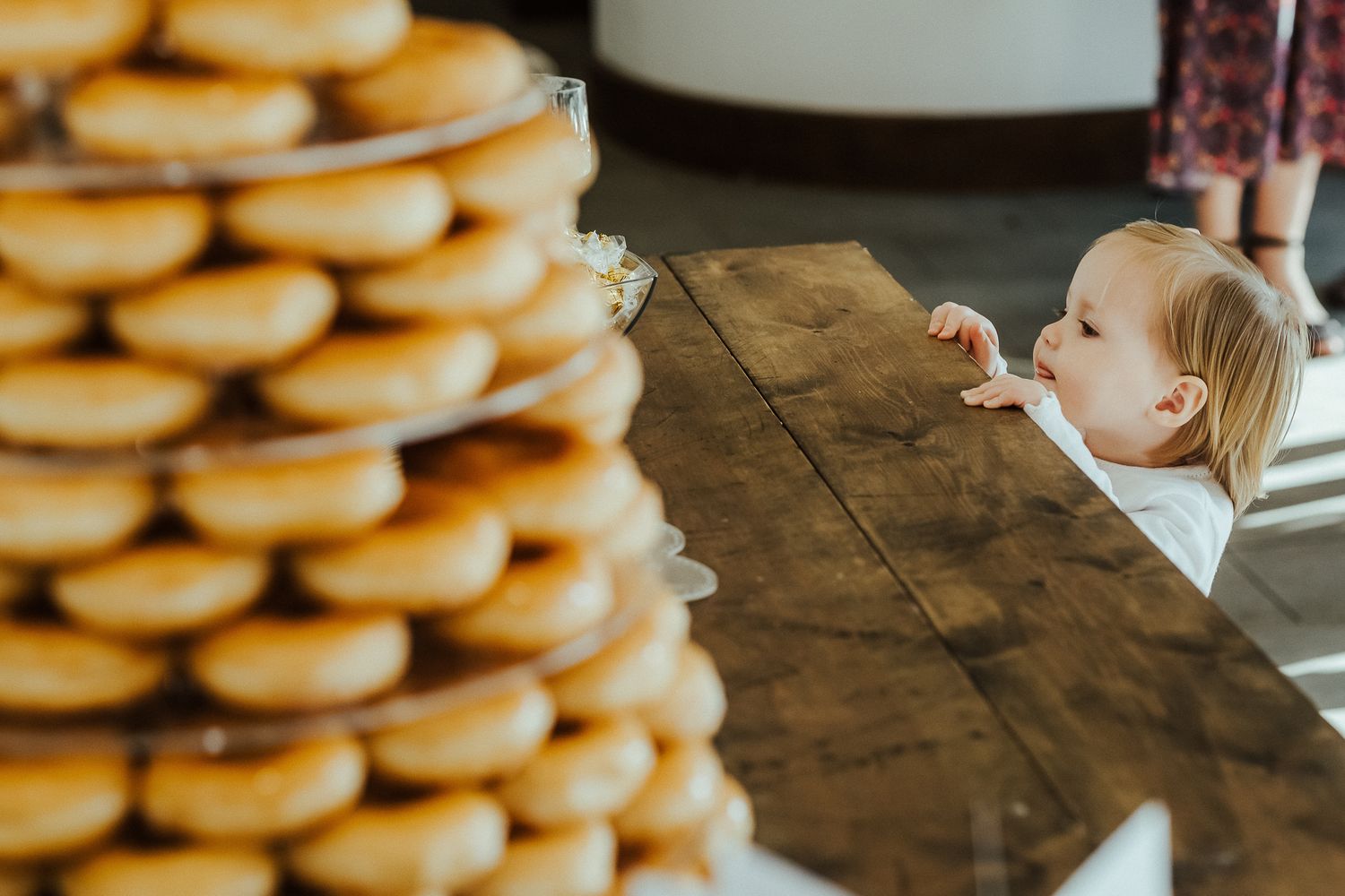 Small child reaches up towards a tall tower of donuts on a wooden table.