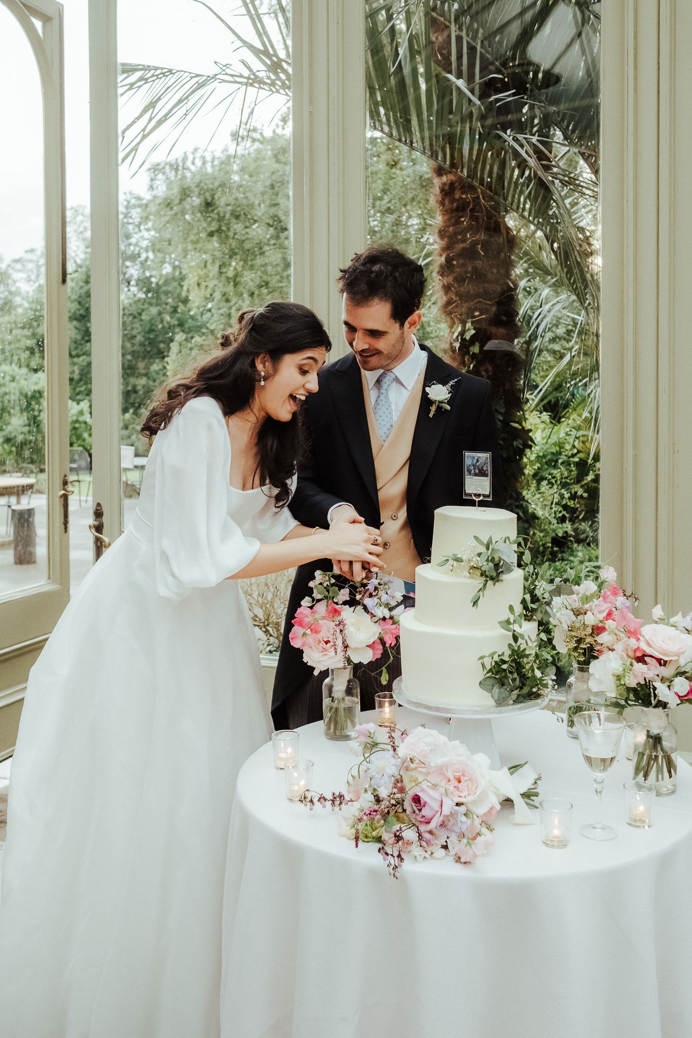 Couple sharing a joyful moment while cutting their wedding cake decorated with pink flowers.