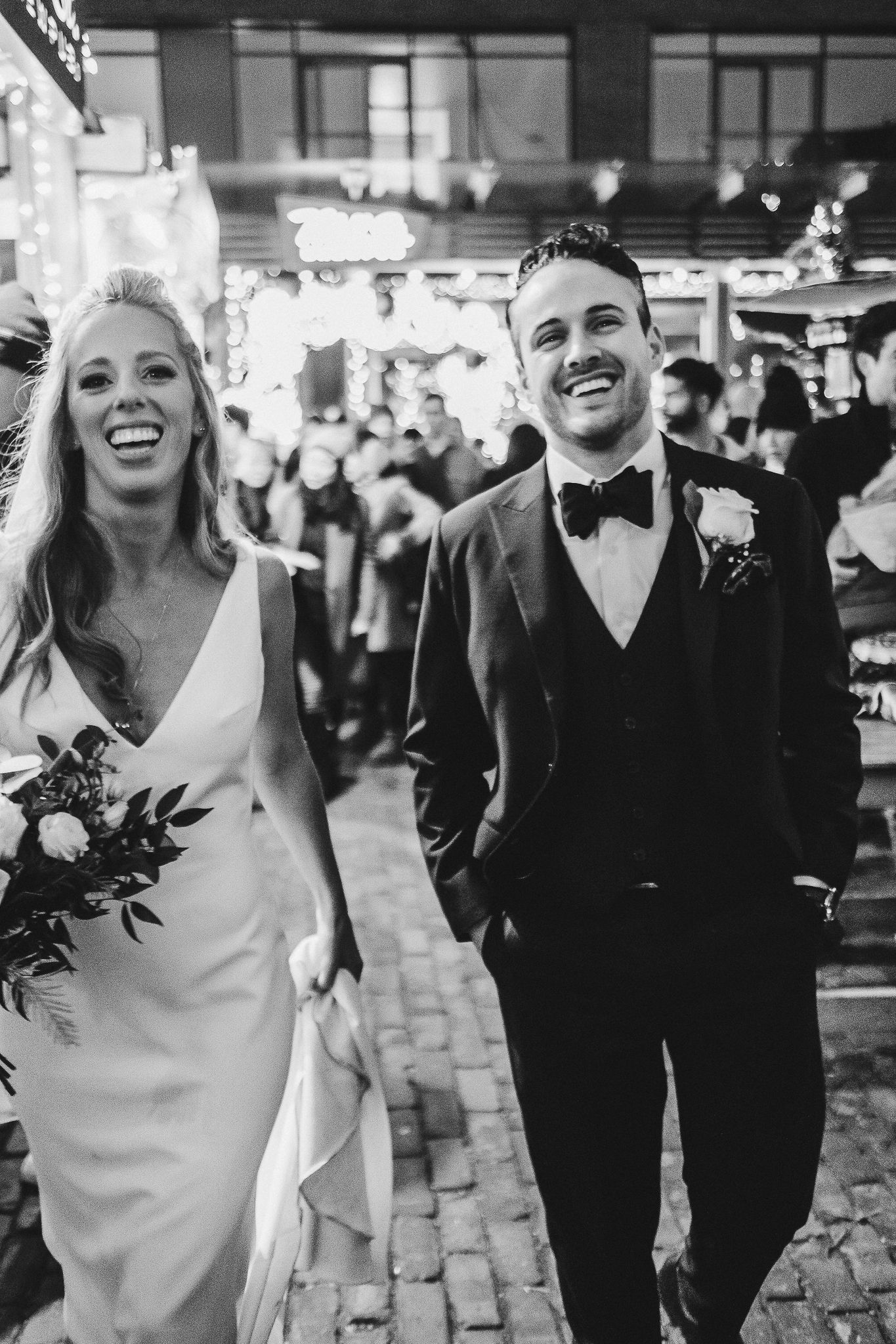 Black and white candid shot of couple walking on cobblestone street with festive lights.