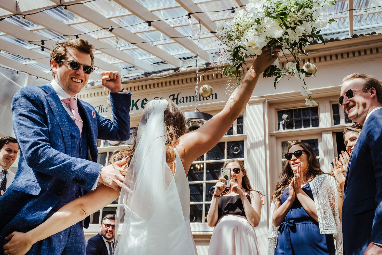 A celebratory moment at an outdoor wedding ceremony under a glass-roofed venue with floral decorations.