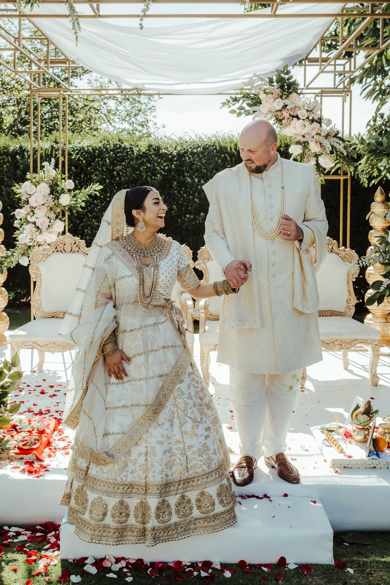 A couple wearing traditional ivory and gold South Asian wedding attire stand together under a canopy during their ceremony.