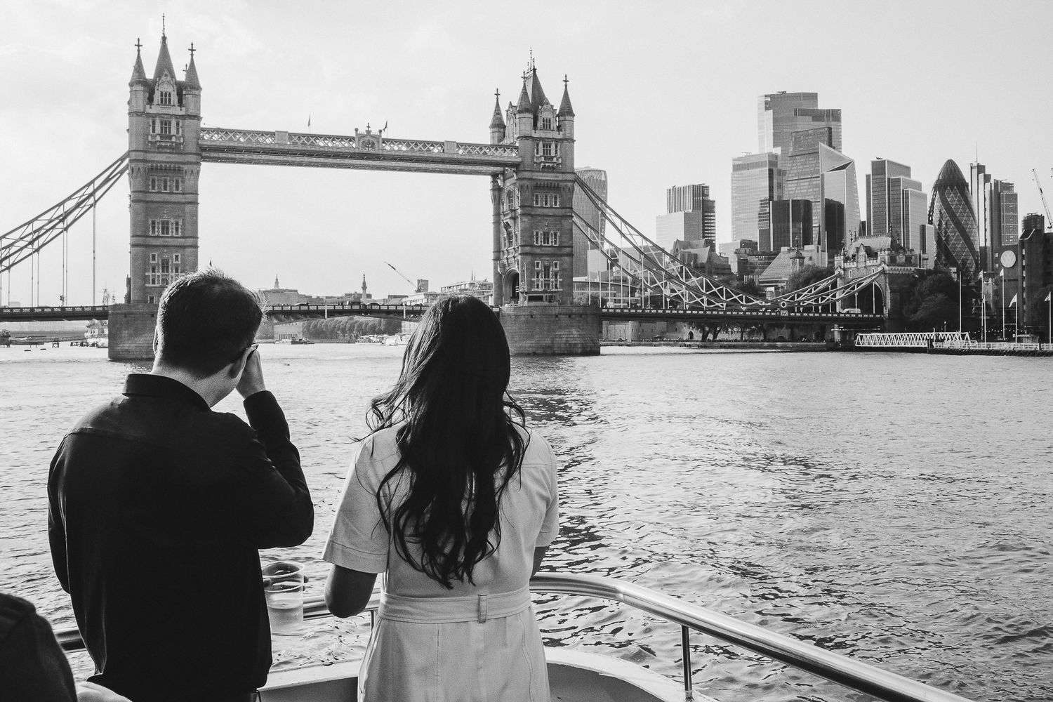 Couple admiring London's Tower Bridge and skyline view from riverside in monochrome photography.