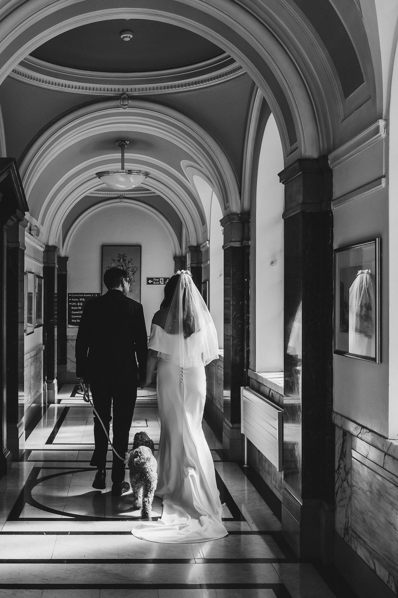 Elegant couple and their dog walk down a grand hallway with arched ceilings and columns in black and white photography.