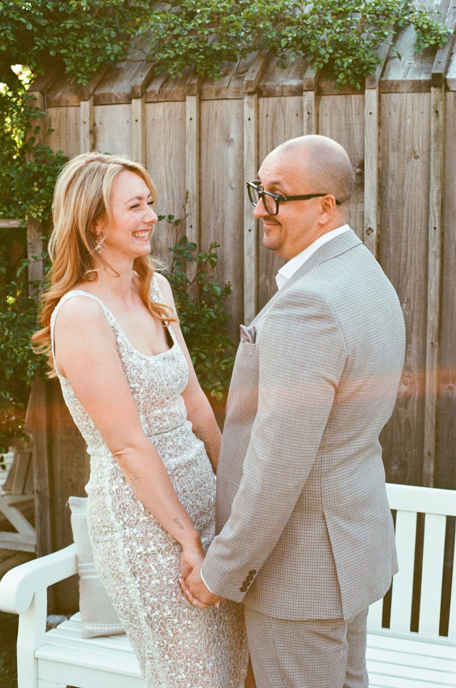 A couple shares laughter and joy while sitting together on a white wooden bench in a garden setting.