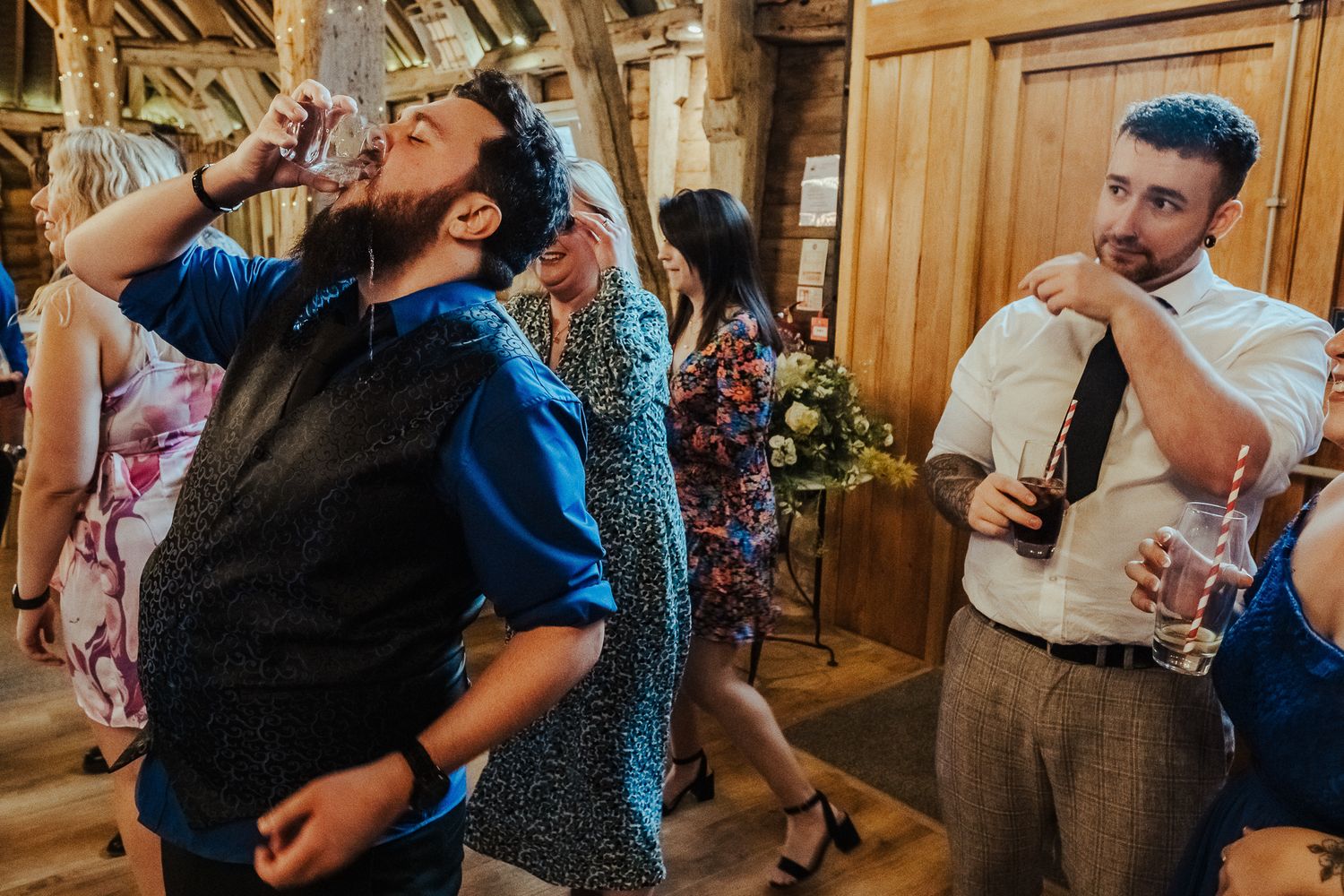 Guest takes dramatic drink while others watch during rustic barn reception.