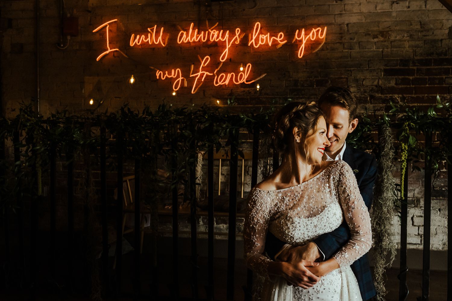 Bride poses beneath glowing neon sign reading I will always love you against brick wall.