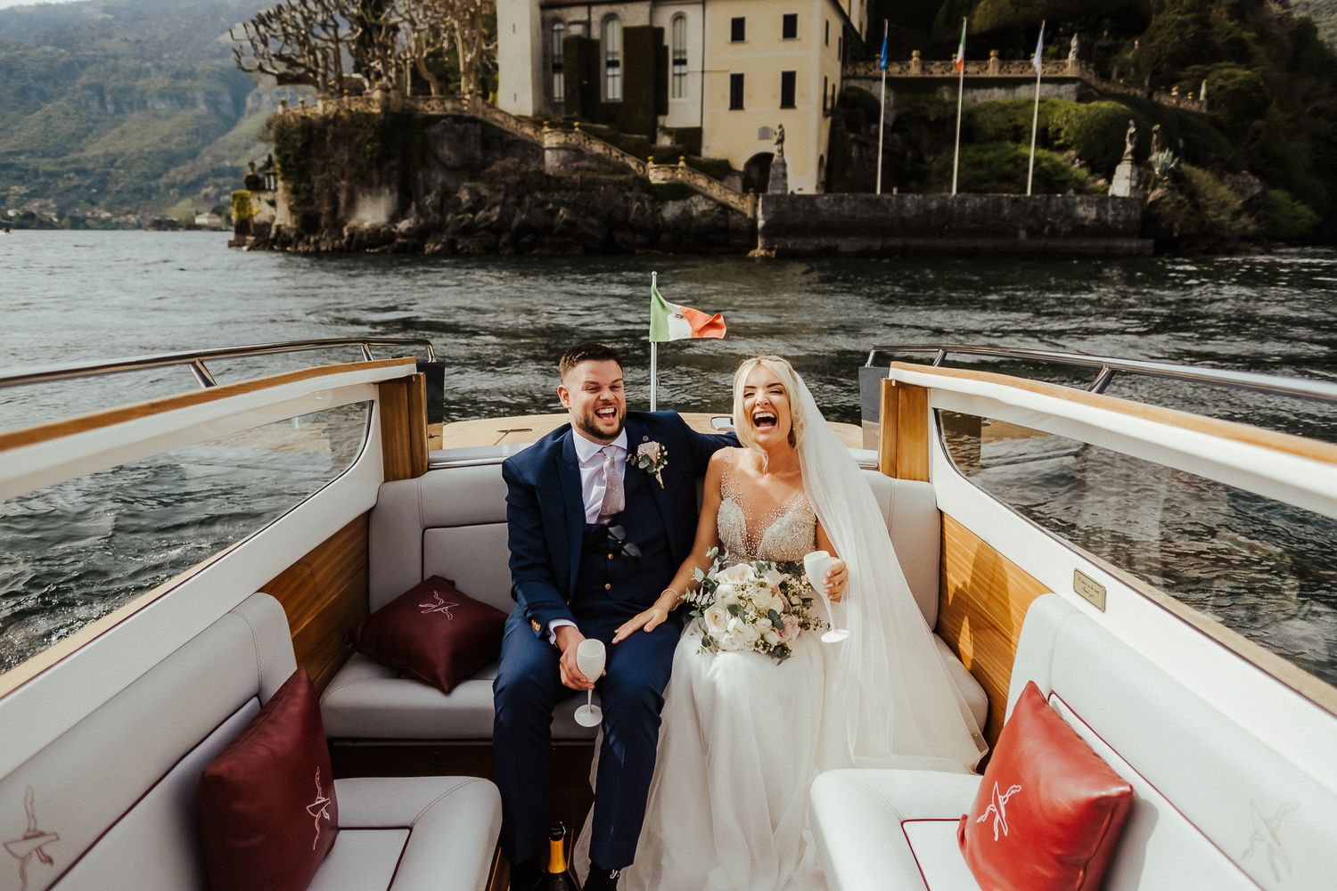 Wedding couple seated in classic wooden boat against Italian lakeside villa backdrop.