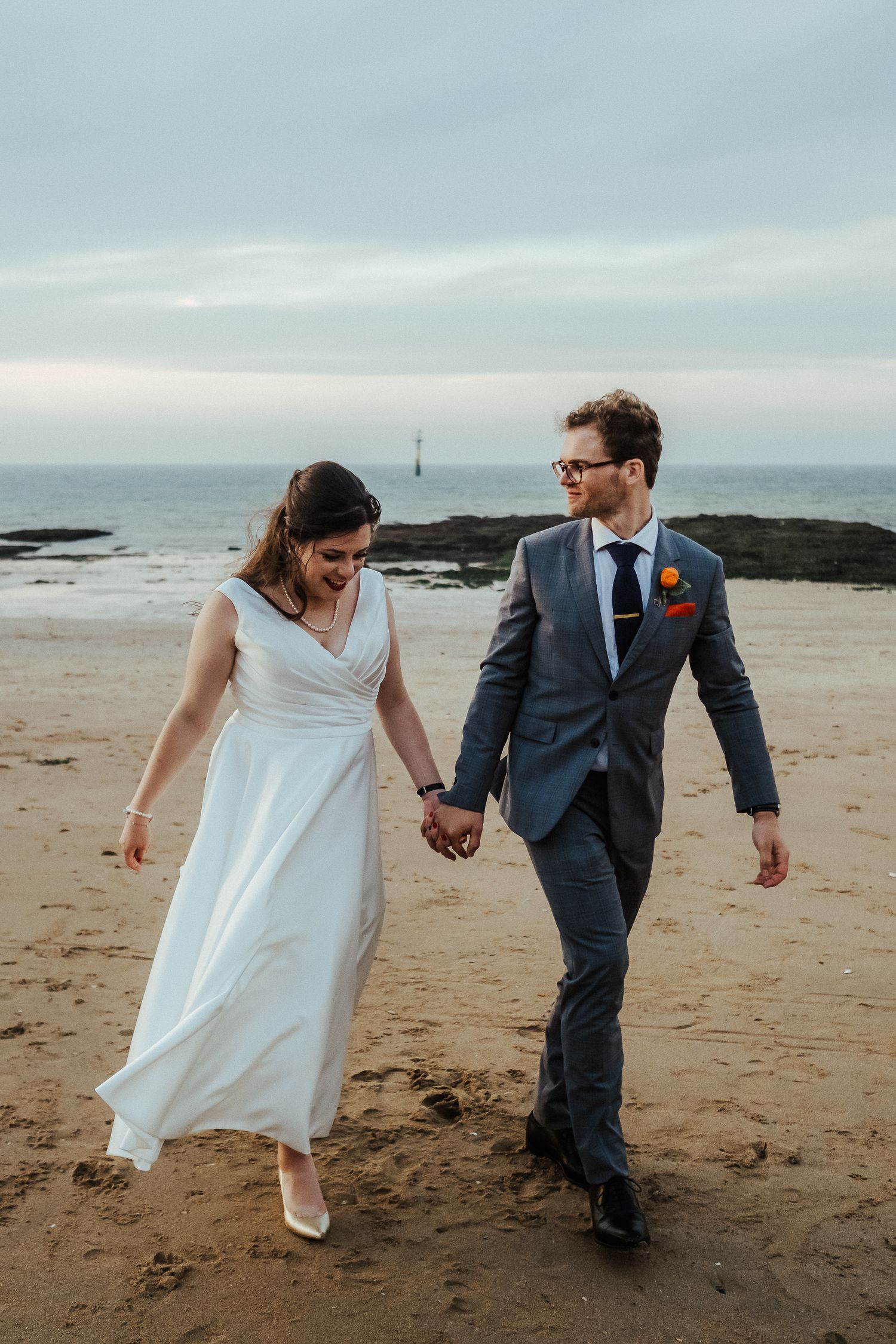 Newlywed couple walking playfully along a rocky beach coastline at sunset.