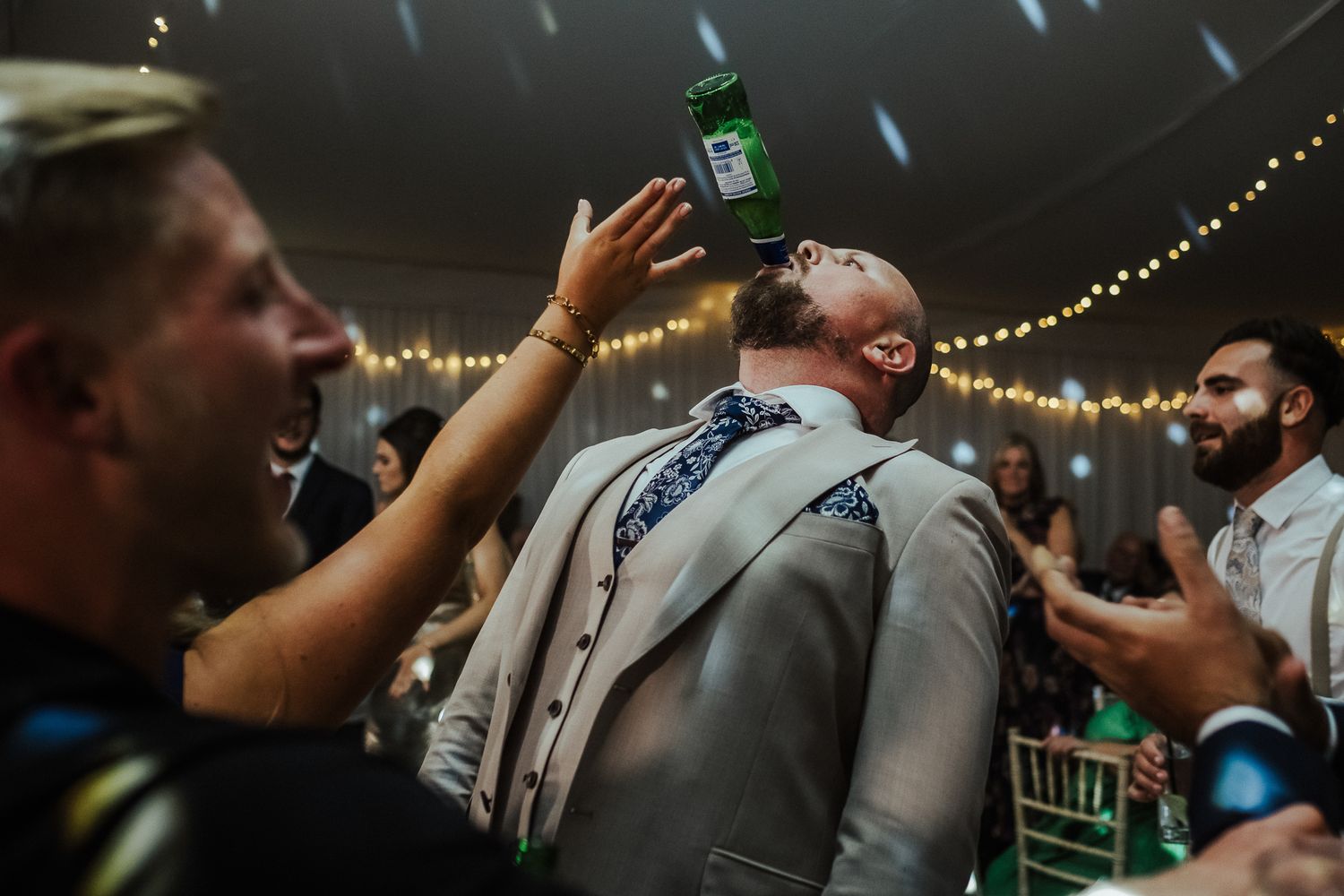 Wedding guests celebrate on dance floor under string lights as someone pours a drink during reception party celebration.