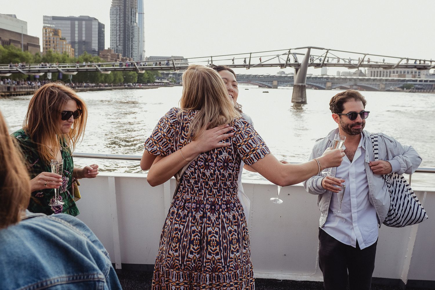 Group of people enjoying drinks on a boat deck with city waterfront and bridge visible in the background.