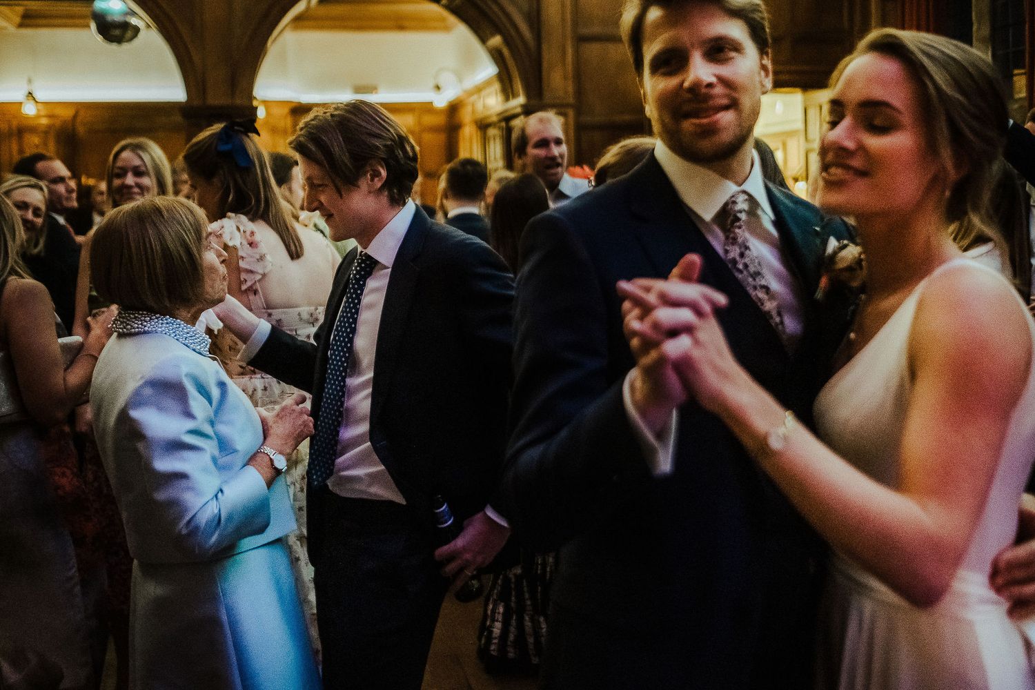 Wedding guests dance together during reception celebrations in an ornate venue with arched doorways.