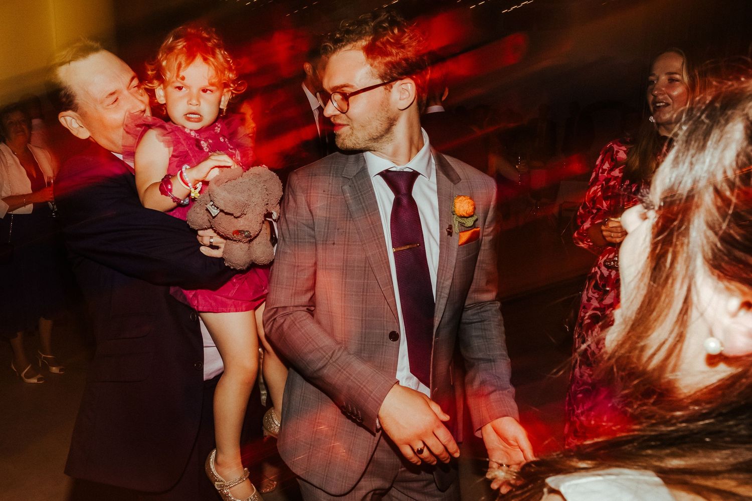 Wedding guest dances with small child under red party lights at reception.