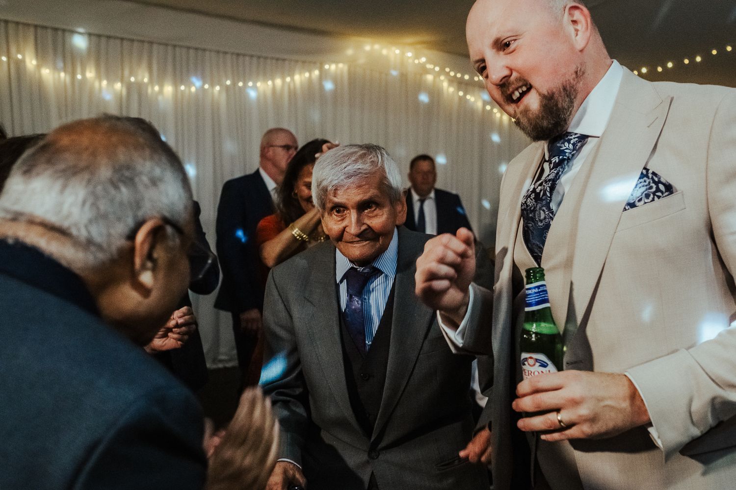Wedding guests dance and celebrate together under twinkling string lights at an evening reception.