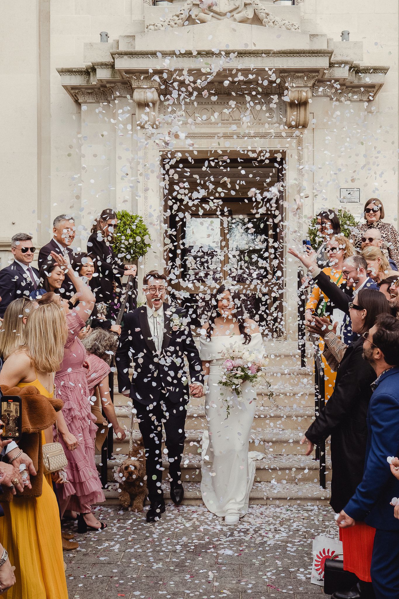 Wedding guests toss confetti in celebration outside a historic building with white stone walls.
