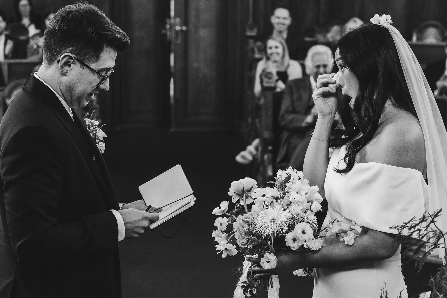 A couple exchanges wedding vows in a church as guests look on in black and white photography.
