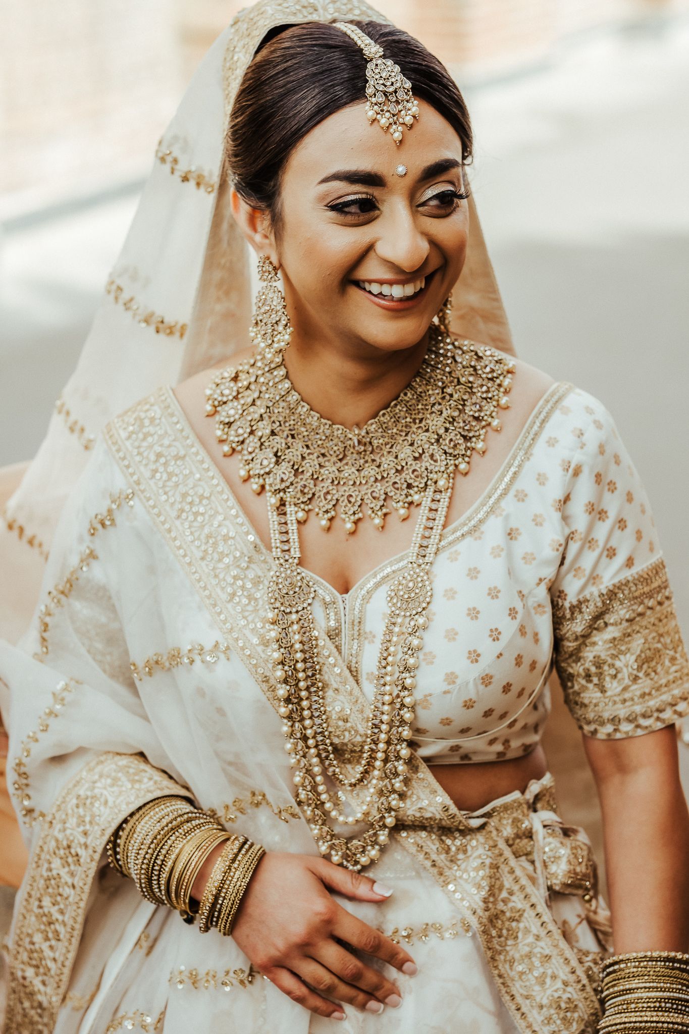 Traditional Indian bride wearing ornate gold jewelry and white embroidered lehenga with matching veil and maang tikka headpiece.