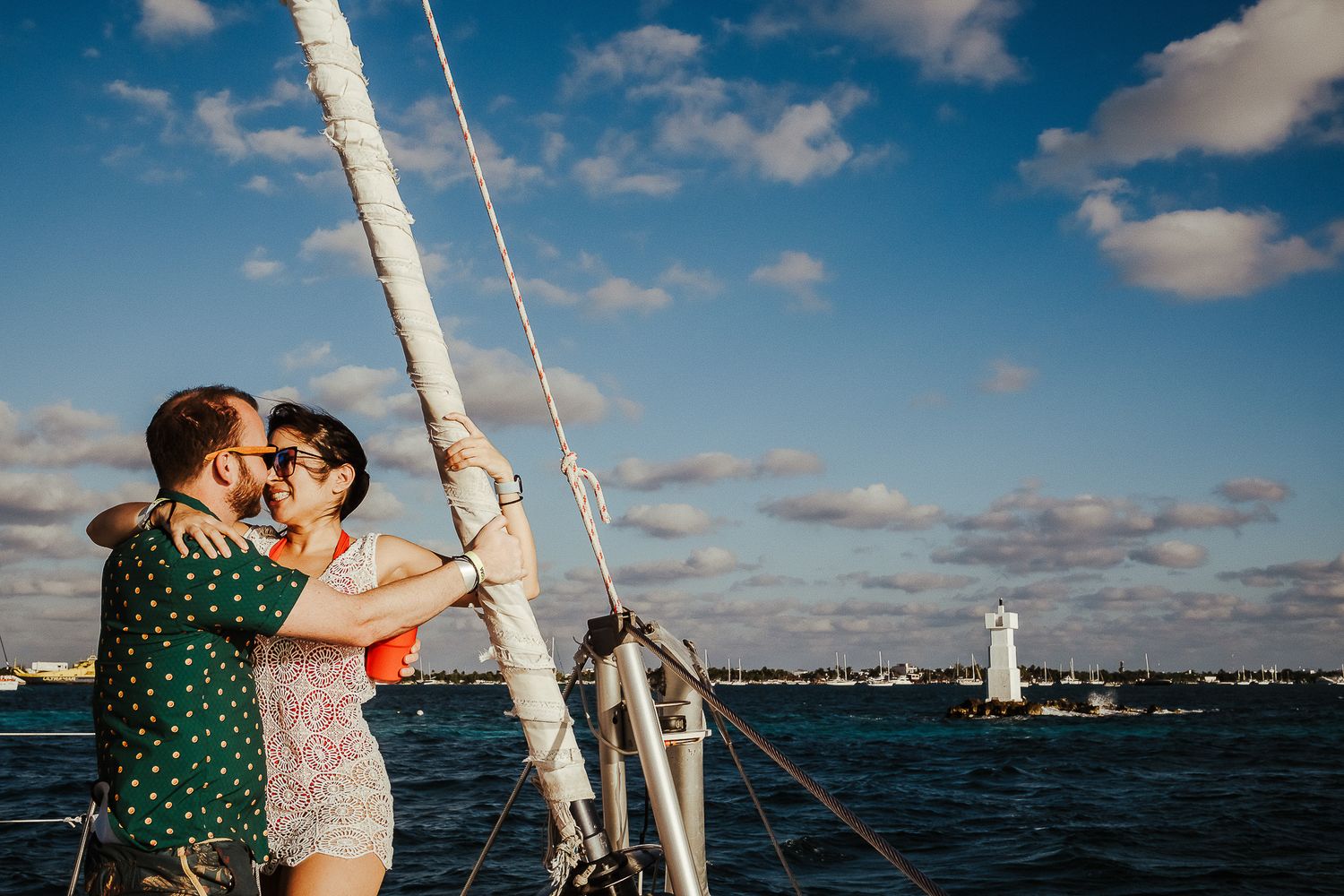 Two people in casual attire sharing an intimate moment on a sailboat with blue skies and water.