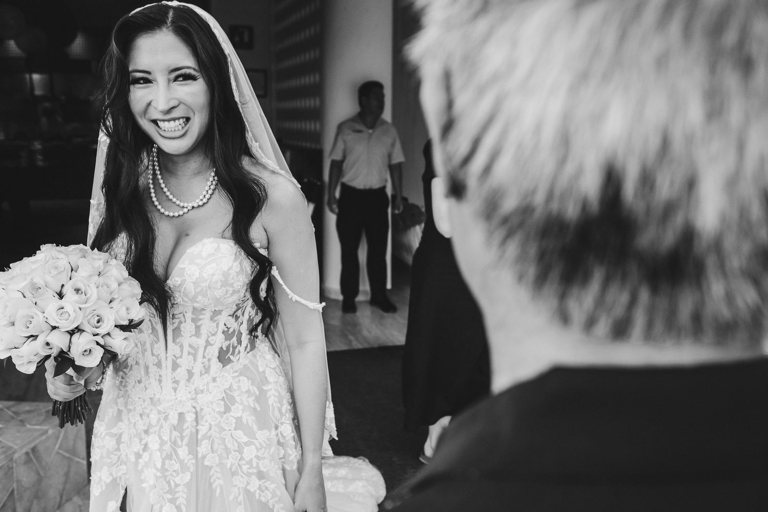 A bride in a white lace dress holds a bouquet of white flowers in an indoor setting.