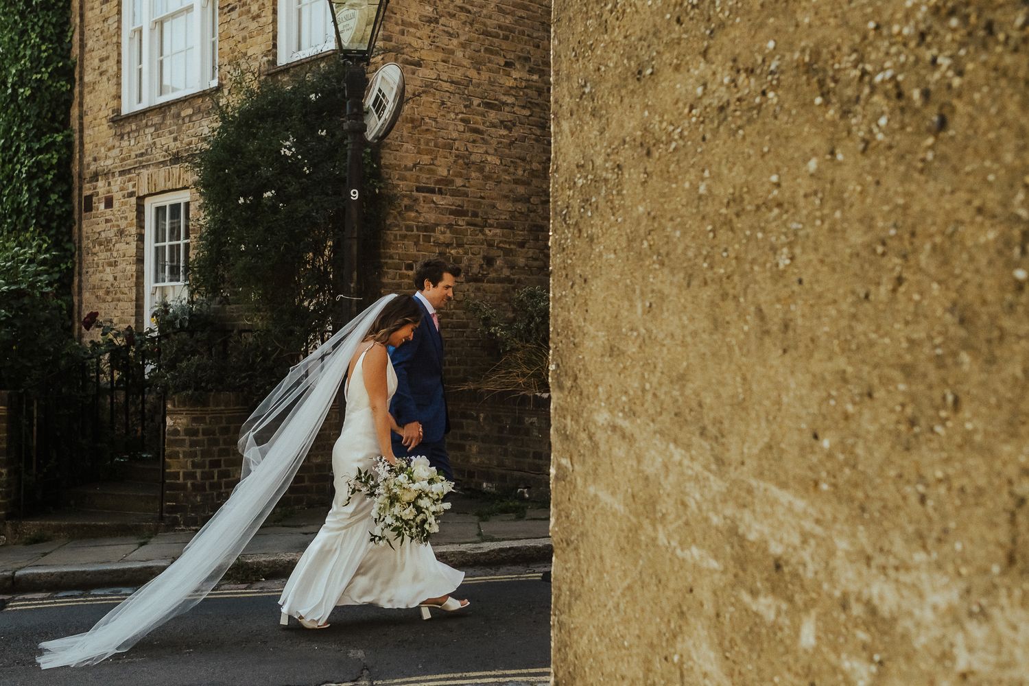 A flowing veil catches the light as a bride walks past a historic stone wall.