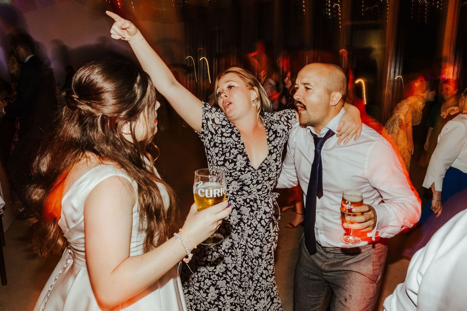 Wedding guests dance enthusiastically on the reception dance floor while holding cocktails.