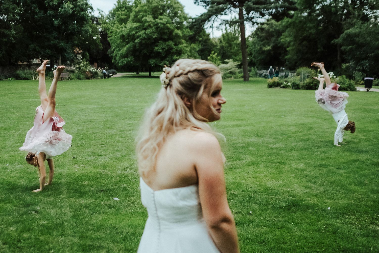 Bride in white dress watches flower girls playing on grassy lawn during outdoor celebration.